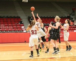 Girls' basketball game: a white-jersey player with number 33 shoots near the basket while black-jersey defenders and teammates watch nearby in a gym with red bleachers behind.