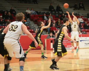 Basketball game action: a white-team player wearing number 2 shoots or passes as defenders in black and yellow close in; spectators sit in red bleachers in the background.