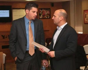 Two businessmen in suits converse in a hallway, one holding a folder or document.