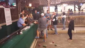 Group of adults socializing at an outdoor bar while playing bocce; colorful bocce balls scattered on the ground around them.