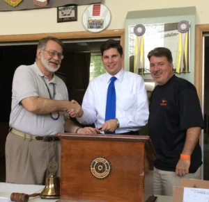 Three men shake hands at a Rotary International podium during a ceremony, with a Rotary emblem on the podium and a bell and gavel on the table.