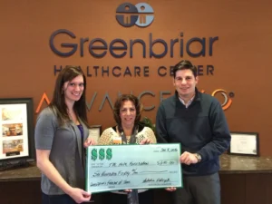 Three people stand in a lobby in front of a wall with the Greenbriar Healthcare Center sign, holding a large ceremonial check.