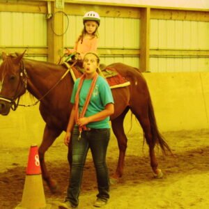 A girl riding a horse indoors while a girl leads the horse