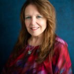 Portrait of a smiling middle-aged woman with long light brown hair wearing a red-purple blouse against a textured blue background.