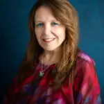 Portrait of a smiling middle-aged woman with long light brown hair wearing a red-purple blouse against a textured blue background.