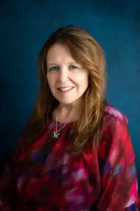 Portrait of a smiling middle-aged woman with long light brown hair wearing a red-purple blouse against a textured blue background.