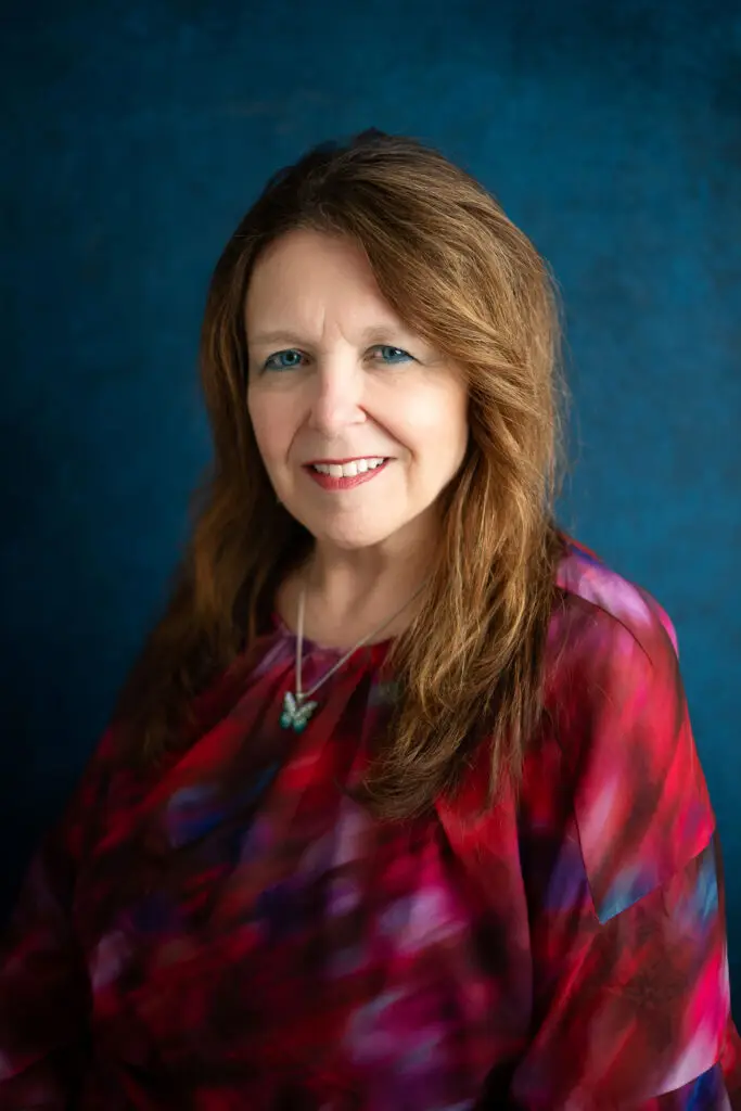 Portrait of a smiling middle-aged woman with long light brown hair wearing a red-purple blouse against a textured blue background.